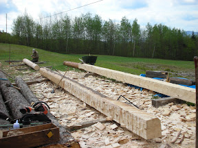 The finished sill timber is in the background with a new sill being roughed out in the foreground.  Michael begins laying out a post timber from a log.