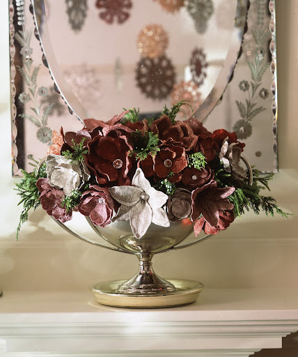 Silk magnolias and lilies are flush with finery: their centers have been replaced with rhinestones, and the petals are coated in red, pink, gold, and silver powder glitters. Real juniper berry branches offer vibrant contrast, and a silver compote provides a fitting base. Silk magnolias and lilies are flush with finery: their centers have been replaced with rhinestones, and the petals are coated in red, pink, gold, and silver powder glitters. Real juniper berry branches offer vibrant contrast, and a silver compote provides a fitting base.