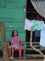 Date: January 2004

Location: Roatan Island, Honduras

Old lady sitting outside her daughters house.  The daughter is inside the house.  Her mother (?) is sitting outside, obviously not in a ggod state.  There is a barbed wire fence in front of her.  Vivid colours.