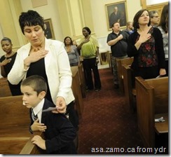 Alina Simona Crehul, left, and her 6-year-old son, Radu, join others in saying the Pledge of Allegiance after she had just taken the naturalization oath with her husband and 23 other new Americans Thursday in the York County Administrative Center. (DAILY RECORD/SUNDAY NEWS--JASON PLOTKIN)