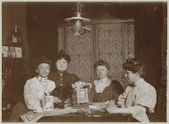 Vier vrouwen aan een tafel met daarop twee fotoportretten en een kat