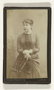 Studioportret van een jonge vrouw in hooggesloten jurk met kantenkraag, leunend tegen een stoel