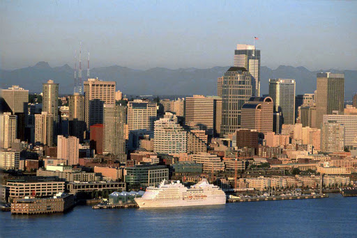 A  cruise ship waits at the Bell Street Pier Cruise Terminal, with the Cascade Mountains seen to the west of the Seattle skyline and Elliott Bay.
