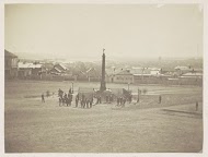 Mensen rondom een zuil met daarop het beeld van een engel, op de achtergrond een stad aan het water