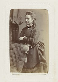 Studioportret van een staande jonge vrouw in een zwarte jurk naast een stoelleuning.