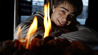 image shows Rybak sitting with his head resting on his arms, with the flames of a fire in the foreground