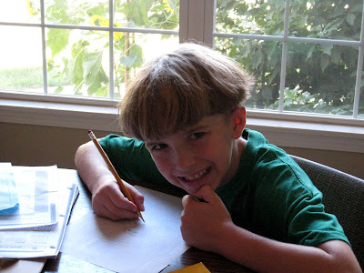 A young boy sitting at a table in front of a window with his homework.