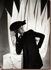 Model, leaning on a square pedestal with white drapery hanging from poles behind her, wearing a dark wool, long jacketed suit with large, figurine buttons, by Schiaparelli, and a black Merry Widow felt hat, inspired by the costumes of Mae West's new film