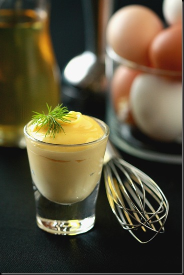 Homemade mayonnaise in a shot glass topped with greens and a whisk with eggs and oil in the background.