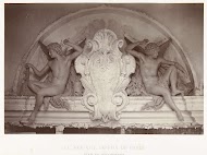 Twee beelden van mannelijke engelen met een hamer in de hand, zittend aan weerszijden van een medaillon, gesitueerd boven een deurpartij.